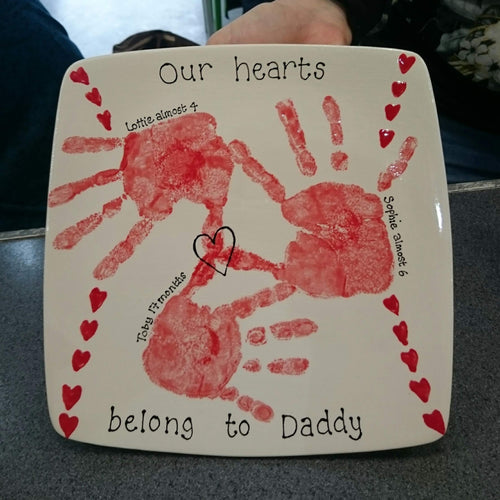 Square plate with three red hand prints and heart decoration with message to Daddy.