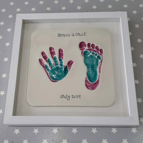 Square Clay Imprint with small hand and footprint inside older siblings hand and footprint. Small prints in teal and larger prints in purple, with white backboard and white frame.