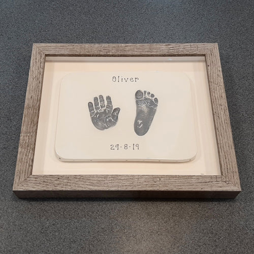 Square Clay Imprint with hand and foot in grey with off white backboard and wooden frame.