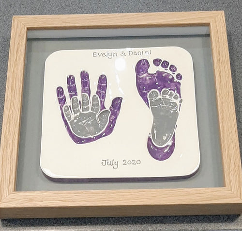 Square Clay Imprint with small hand and footprint impressions inside older siblings hand and footprint impressions.  Larger prints in purple and smaller prints in grey with grey back board and wooden frame.