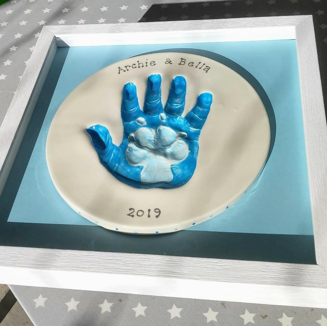 Circle Clay Imprint with paw print inside hand print.  Paw print in pale blue and hand print in a darker blue with blue backboard and white frame.