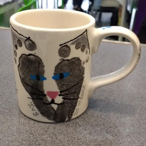 Two black footprints on a Regular Mug making the shape of a cat's face with eyes, nose and whiskers added to complete the face.