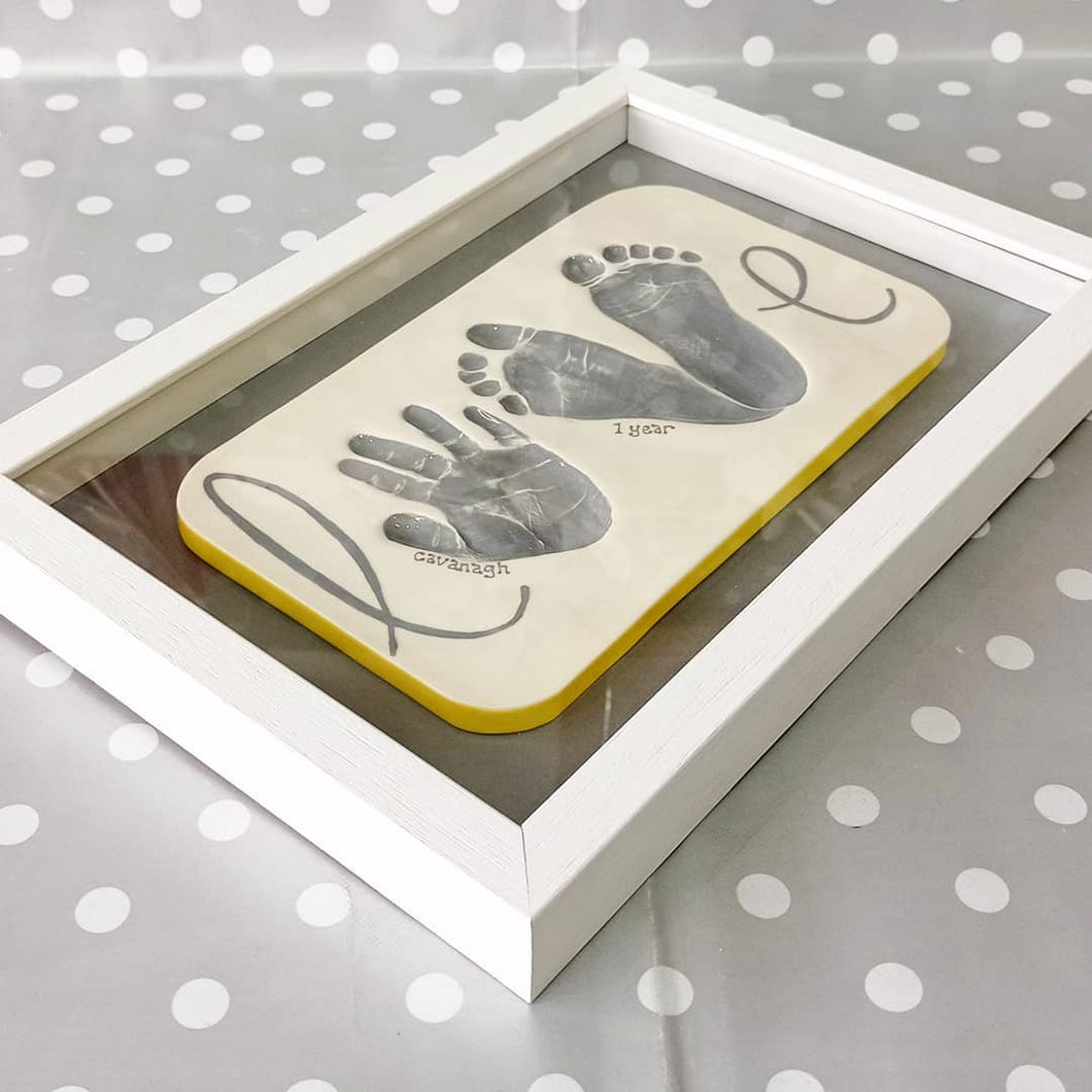 A clay imprint spelling the word LOVE with a hand print and two footprints, painted in grey with a white frame.