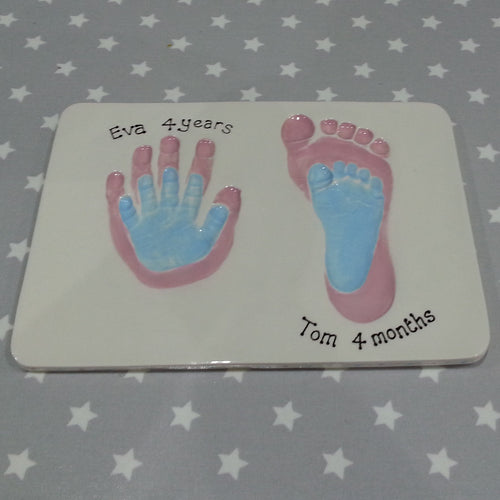 Rectangle Clay Imprint with small hand and foot prints inside older siblings hand and footprint.  Smaller prints in pastel blue and larger prints in pink.  Unframed. 