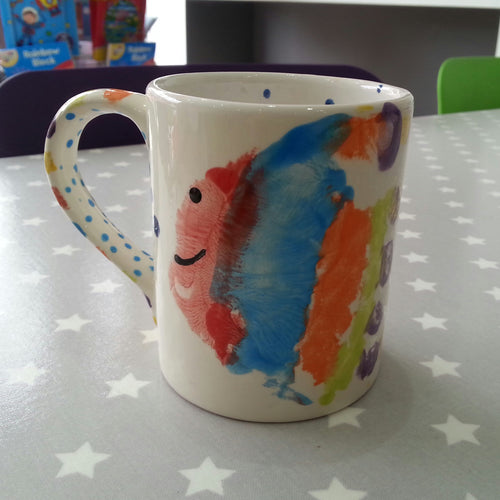 Multi coloured hand print turned into a rainbow fish on a regular mug, decorated with dots.
