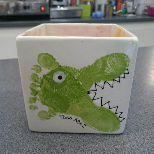 Square Herb Pot with two green footprints, positioned in such a way to be turned into a crocodile.  Teeth and eye added to create the transformation. 