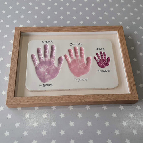 Rectangle Clay Imprint with three sibling hand prints in lilac, pink and purple with off white back board and oak frame.