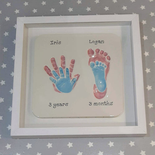 Square Clay Imprint with small hand a footprint impression inside older siblings hand and footprint impression.  Larger prints in pastel pink, smaller prints in pastel blue with white backboard and white frame.