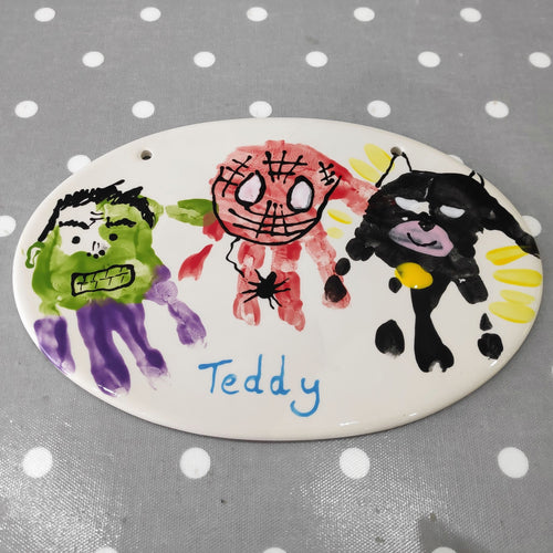 An Oval Plaque with purple and green hand print turned into the Hulk, red hand print turned into Spiderman and black hand print turned into Batman.