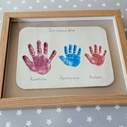 Rectangle Clay Imprint with three sibling hand prints in purple, blue and red.  Beige backboard and wooden frame. 
