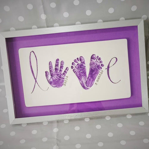 LOVE Clay Imprint using a hand and two footprints to spell the work LOVE. Purple prints with purple backboard and silver frame.
